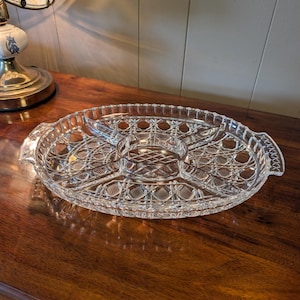 Peut inclure: Un plateau de service en verre transparent avec un design divisé comprenant un compartiment circulaire central entouré de quatre sections extérieures. Le plateau présente un motif en losanges texturé, un bord festonné et deux poignées intégrées.