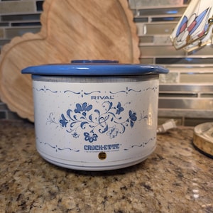 May include: A white Crock-Ette slow cooker with a blue lid and floral design. The slow cooker has the brand name "Rival" and the text "Crock-Ette 1 Qt. Stoneware Slow Cooker" printed on the front.