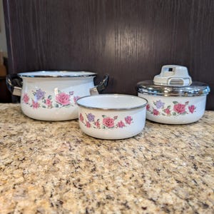 Puede incluir: Un juego de tres recipientes de cocina de esmalte blanco con un patrón floral de rosas rosas y moradas. El juego incluye una olla grande con dos asas laterales negras, un cuenco mediano y un recipiente más pequeño con tapa y borde plateado.