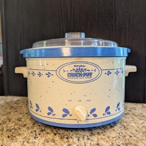 May include: A Rival Crock-Pot stoneware slow cooker with a cream-colored base featuring blue speckles and a blue leaf pattern. It has a clear glass lid, two side handles, and a front control knob with settings for off, low, and high heat.