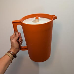 May include: A bright orange plastic pitcher featuring a tall cylindrical body, a sturdy side handle, and a white circular lid with a small orange center knob. The pitcher includes a molded pouring spout at the top rim for liquid dispensing.
