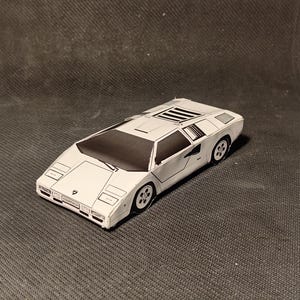 May include: A white toy car with black accents, resembling a classic sports car. The model has detailed features, including wheels, windows, and a distinctive wedge shape. The car is positioned on a dark gray surface.