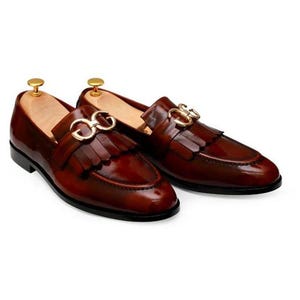 Puede incluir: Un par de mocasines de cuero burdeos pulido con herrajes dorados y flecos. Los zapatos tienen un dise&ntilde;o cl&aacute;sico sin cordones con un tac&oacute;n bajo y un acabado elegante y brillante. Los mocasines se muestran sobre un fondo blanco.