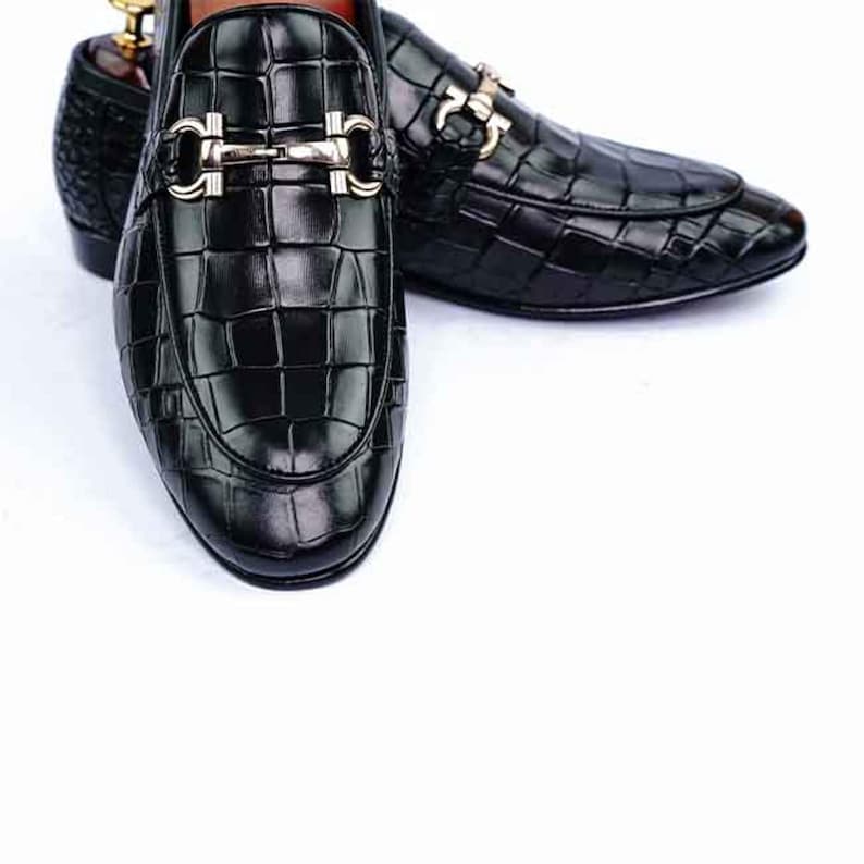 May include: Black leather loafers with a crocodile texture. The shoes have a silver-colored buckle and a rounded toe. The loafers are displayed on a white background. The shoes are a classic style.