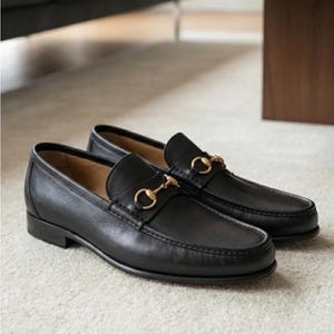 Könnte beinhalten: Ein Paar schwarze Leder-Loafer mit goldfarbenen Metall-Horsebit-Details. Die Schuhe werden auf einem hellen Teppich präsentiert, der ihr klassisches Design und ihr poliertes Aussehen zeigt. Die Loafer haben einen niedrigen Absatz und eine runde Spitze.