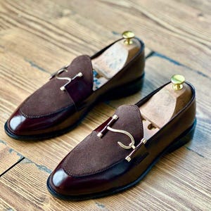 Puede incluir: Un par de mocasines de cuero burdeos con detalles de ante y herrajes dorados. Los zapatos tienen un dise&ntilde;o cl&aacute;sico con un acabado pulido y se exhiben sobre una superficie de madera.