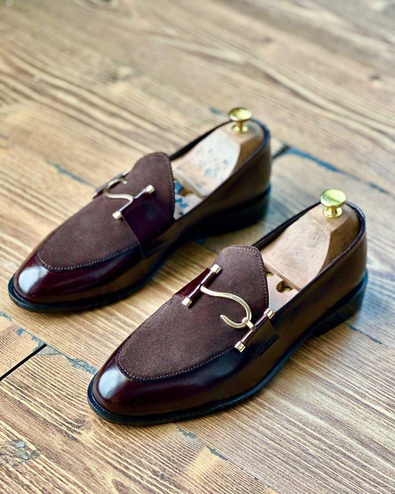 Puede incluir: Un par de mocasines de cuero burdeos con detalles de ante y herrajes dorados. Los zapatos tienen un dise&ntilde;o cl&aacute;sico con un acabado pulido y se exhiben sobre una superficie de madera.