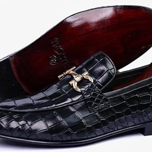 May include: Black leather loafers with a crocodile skin texture. The shoes have a gold-colored buckle and a dark brown sole. One shoe is upright, and the other is upside down, showing the sole.
