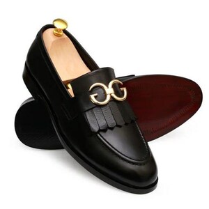 Puede incluir: Zapatos mocasines de cuero negro con hebilla dorada y detalle de flecos. Los zapatos tienen un dise&ntilde;o cl&aacute;sico con punta redondeada y suela marr&oacute;n oscuro. Los zapatos se muestran sobre un fondo blanco.