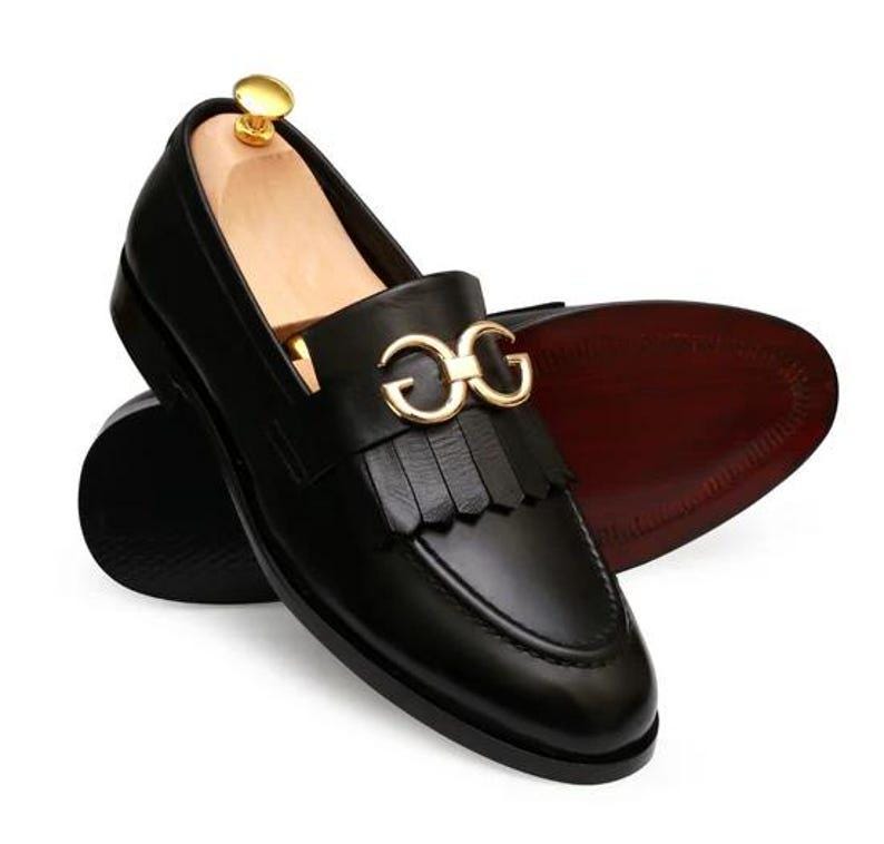 Puede incluir: Zapatos mocasines de cuero negro con hebilla dorada y detalle de flecos. Los zapatos tienen un dise&ntilde;o cl&aacute;sico con punta redondeada y suela marr&oacute;n oscuro. Los zapatos se muestran sobre un fondo blanco.