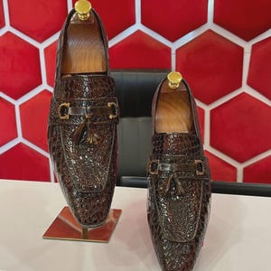 以下が含まれることがあります： クロコダイル柄の茶色の革製ローファー2足。各靴には金色のバックルとタッセルのディテールがあります。靴は木製のシューツリーにセットされ、白い表面に展示されています。
