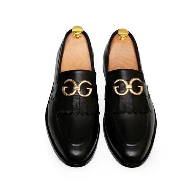 Puede incluir: Un par de mocasines de cuero negro con herrajes dorados. Los zapatos presentan una hebilla dorada decorativa y detalles de flecos. Los mocasines se exhiben sobre hormas de madera sobre un fondo blanco.