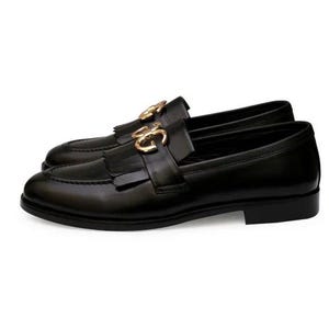 Puede incluir: Un par de mocasines de cuero negro con herrajes dorados. Los zapatos presentan un dise&ntilde;o cl&aacute;sico con punta redondeada, una correa decorativa con hebilla dorada y borlas. Talla 43 (EU).