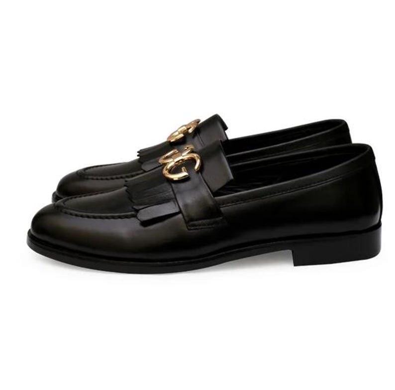 Puede incluir: Un par de mocasines de cuero negro con herrajes dorados. Los zapatos presentan un dise&ntilde;o cl&aacute;sico con punta redondeada, una correa decorativa con hebilla dorada y borlas. Talla 43 (EU).