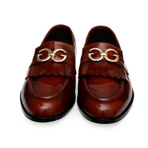 Puede incluir: Un par de mocasines de cuero marr&oacute;n con un acabado brillante. Cada zapato presenta una hebilla dorada y un detalle de flecos en el empeine. Los zapatos tienen un aspecto cl&aacute;sico y pulido.