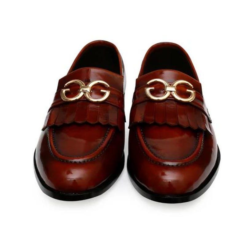 Puede incluir: Un par de mocasines de cuero marr&oacute;n con un acabado brillante. Cada zapato presenta una hebilla dorada y un detalle de flecos en el empeine. Los zapatos tienen un aspecto cl&aacute;sico y pulido.