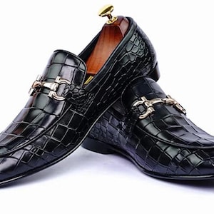 May include: A pair of black leather loafers with a crocodile texture. The shoes feature a gold-colored buckle and a wooden shoe tree inside. The loafers are designed for formal or business attire.