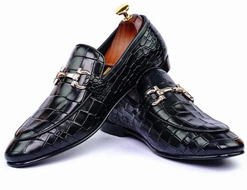 May include: A pair of black leather loafers with a crocodile texture. The shoes feature a gold-colored buckle and a wooden shoe tree inside. The loafers are designed for formal or business attire.