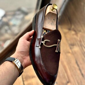 Puede incluir: Un mocas&iacute;n de cuero marr&oacute;n con empeine de ante y herrajes dorados. El zapato presenta un dise&ntilde;o cl&aacute;sico con un acabado pulido. El zapato est&aacute; sostenido por una persona.