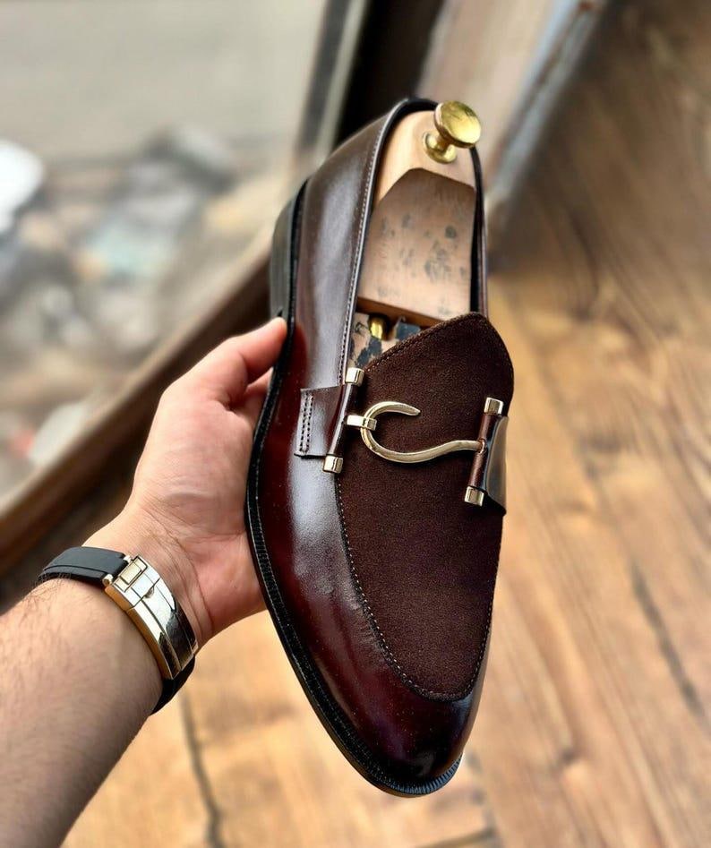 Puede incluir: Un mocas&iacute;n de cuero marr&oacute;n con empeine de ante y herrajes dorados. El zapato presenta un dise&ntilde;o cl&aacute;sico con un acabado pulido. El zapato est&aacute; sostenido por una persona.