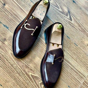 Puede incluir: Un par de mocasines de cuero marr&oacute;n con detalles de ante y herrajes dorados. Los zapatos tienen un acabado pulido y se exhiben sobre una superficie de madera. Se insertan hormas dentro de los zapatos.