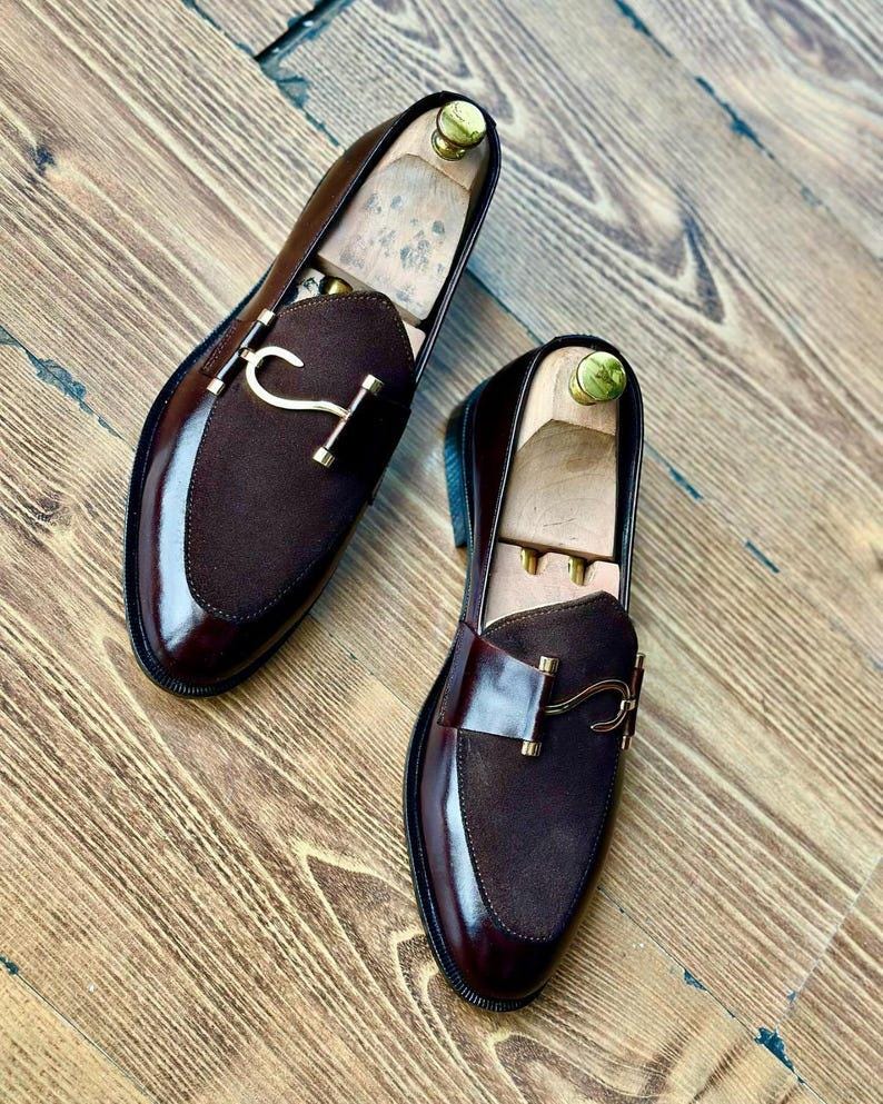 Puede incluir: Un par de mocasines de cuero marr&oacute;n con detalles de ante y herrajes dorados. Los zapatos tienen un acabado pulido y se exhiben sobre una superficie de madera. Se insertan hormas dentro de los zapatos.