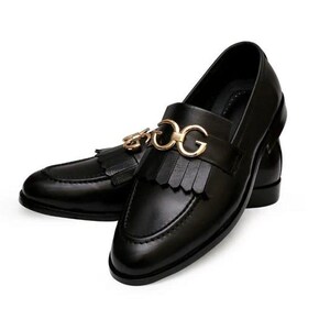 Puede incluir: Un par de mocasines de cuero negro con un adorno met&aacute;lico dorado con las letras "DG". Los zapatos tienen un detalle de borlas y un dise&ntilde;o cl&aacute;sico y pulido, adecuado para ocasiones formales o semiformales.