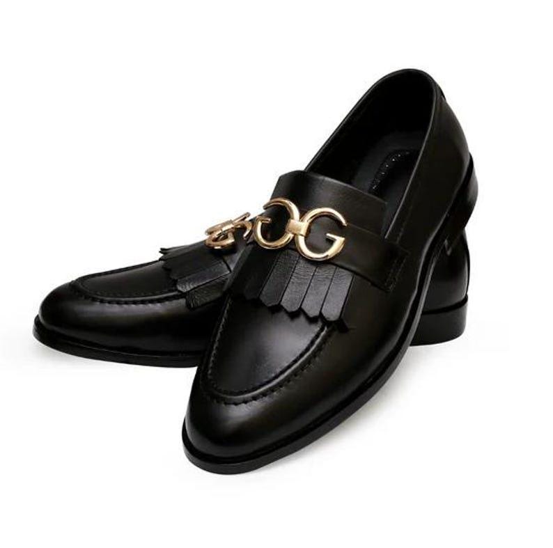 Puede incluir: Un par de mocasines de cuero negro con un adorno met&aacute;lico dorado con las letras "DG". Los zapatos tienen un detalle de borlas y un dise&ntilde;o cl&aacute;sico y pulido, adecuado para ocasiones formales o semiformales.