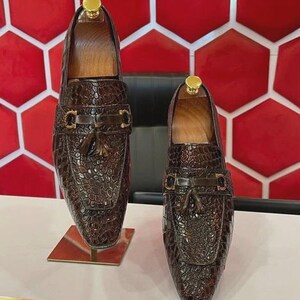 以下が含まれることがあります： クロコダイル柄のブラウンレザーローファー2足。各靴には金色のバックルとタッセルのディテールがあります。靴は、白の表面に、赤と白の幾何学模様の背景で展示されています。