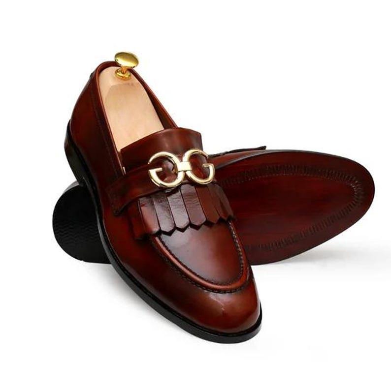 Puede incluir: Mocasines de cuero marr&oacute;n con hebilla dorada y detalles de flecos. Los zapatos tienen un dise&ntilde;o cl&aacute;sico con un acabado pulido, adecuados para ocasiones formales o semiformales. Las suelas son de color marr&oacute;n oscuro.