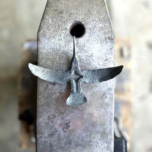 以下が含まれることがあります： 金属製の鳥の彫刻。翼を広げた鳥が、質感のある金属の表面に置かれています。鳥はエッチングされた線で詳細に表現され、職人技を示しています。彫刻の濃いグレーの色は、明るい金属の背景と対照的です。