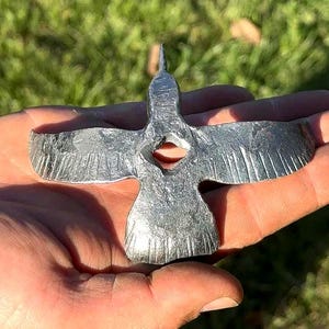 以下が含まれることがあります： 手作りの銀色の金属製の鳥の彫刻で、翼を広げています。鳥は尖ったくちばしと体の中心に穴があります。翼には詳細な羽の模様があります。彫刻は、ぼやけた緑色の背景に対して、手で持たれています。