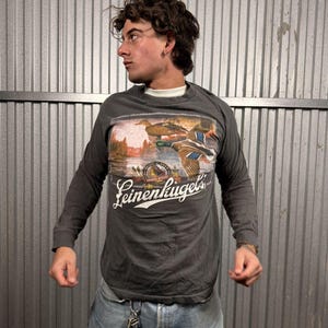 May include: Dark gray long-sleeve t-shirt featuring a graphic of ducks in flight over a lake, with the text "Leinenkugel's" in white script. The shirt is worn with blue jeans and a white undershirt.