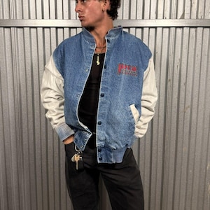Op de afbeelding: Een blauw spijkerjack met witte mouwen en een rode logopatch. De jas heeft een knoopsluiting en geribde manchetten en zoom. De persoon draagt een zwarte broek en een zwart shirt.