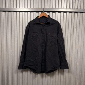 Könnte beinhalten: Schwarzes Langarmhemd mit Knopfleiste, zwei Brusttaschen und Kragen. Es hängt an einem Holzbügel. Das Wrangler-Logo ist sichtbar. Der Hintergrund ist aus Wellblech.