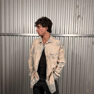 May include: A light-colored, long-sleeved button-down shirt with a faded blue pattern. The shirt has two chest pockets and is worn over a black shirt. The person is wearing black pants and a black belt.