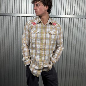 May include: A plaid button-down shirt with a yellow, white, and lavender pattern. The shirt features red rose embroidery on the shoulders and decorative stitching. The shirt has a classic western style with front pockets.