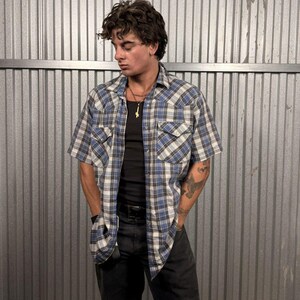 May include: A short-sleeved, blue and white checked shirt with two chest pockets. The shirt is worn over a black vest top and paired with black trousers. A gold cross necklace and a black belt complete the outfit.
