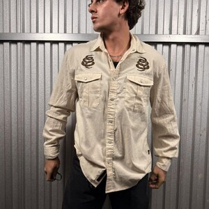 May include: A long-sleeved, cream-colored button-down shirt featuring two chest pockets and a coiled snake graphic embroidered in black and gold on each side of the chest. The shirt has a classic collar and silver-toned metal snap buttons.