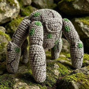 Puede incluir: Una criatura tejida a ganchillo con un cuerpo redondeado y cuatro patas gruesas y robustas. El hilo es de color gris claro con pequeñas manchas verdes dispersas y líneas bordadas en marrón oscuro que imitan la apariencia de piedra agrietada.