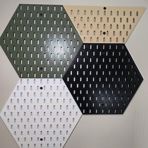 May include: A set of hexagonal pegboards in olive green, beige, black, and white. Each board has a series of evenly spaced holes for hanging items. The boards are mounted on a neutral wall, creating a modern storage solution.
