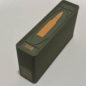 May include: An olive green rectangular box with a raised orange bullet design on the top. The text ".308" is in gold on the top and "308 WIN" is on the side. The box is angled, showing its side and top.