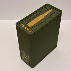 May include: An olive green rectangular box with a hinged lid. The lid has a raised outline of a bullet and the text ".50 BMG" in gold. The side of the box has a gold label with the text ".50 CAL BMG".