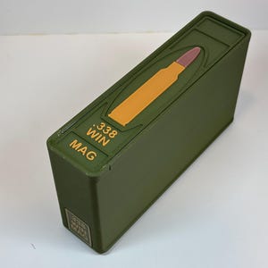 Puede incluir: Una caja de cargador de munición verde oliva con un gráfico de bala amarilla y marrón en la parte superior. La caja tiene el texto ".338 WIN MAG" en amarillo. La caja es rectangular y tiene una tapa con bisagras.