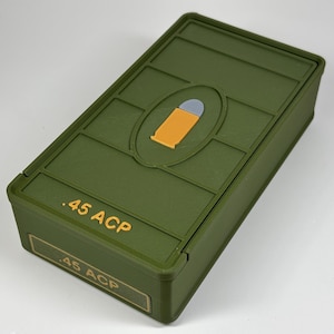 May include: Olive green rectangular box with a hinged lid. The box features a raised oval design with a bullet graphic in orange and gray. The text ".45 ACP" is printed in yellow on the box.