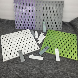 Könnte beinhalten: Eine Auswahl an farbenfrohen Lochplatten und Befestigungsmaterial. Die Platten sind in Weiß, Grün, Grau und schillerndem Lila gehalten. Das Befestigungsmaterial ist in Weiß und Grau gehalten. Die Lochplatten haben gleichmäßig verteilte ovale Löcher.