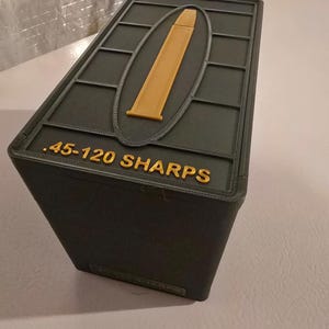 Pode incluir: Uma caixa retangular verde escuro com um desenho de bala dourada na tampa. O texto ".45-120 SHARPS" é exibido em letras douradas. A caixa é provavelmente para armazenar munição.
