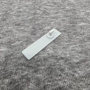 Op de afbeelding: Een lichtgrijs, rechthoekig plastic object met een cirkelvormige knop aan één uiteinde. Het object ligt op een grijze, getextureerde ondergrond. De knop heeft een klein, verhoogd detail.