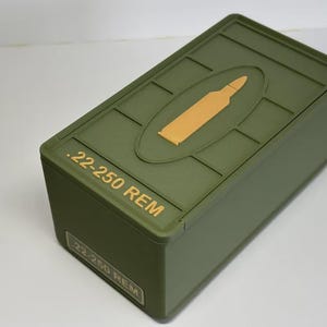 May include: An olive green rectangular box with a hinged lid. The lid features a gold-colored bullet graphic inside an oval and the text ".22-250 REM". The box has a label with the same text.