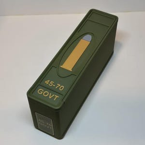 Könnte beinhalten: Eine olivgrüne rechteckige Box mit einer goldfarbenen Geschossgrafik und dem Text "45-70 GOVT". Die Box hat einen Klappdeckel und ein kleines rechteckiges Etikett an der Seite mit dem gleichen Text.